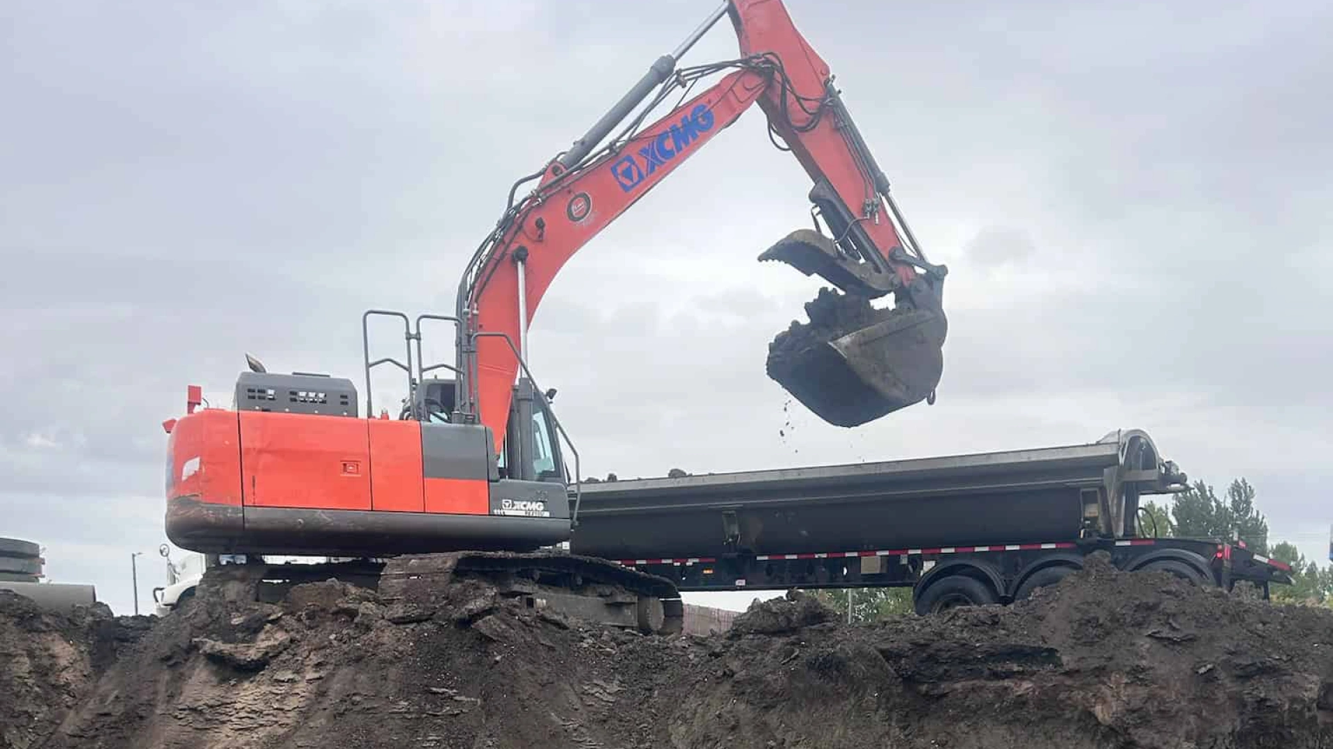 A construction site on an overcast day with an orange excavator dumping dirt into a black trailer A construction site on an overcast day with an orange excavator dumping dirt into a black trailer